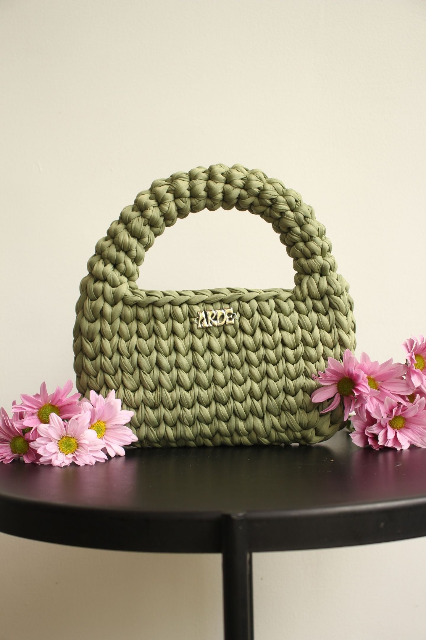 Soft green crocheted handbag with 'ARDE' brand on a black table with pink flowers.