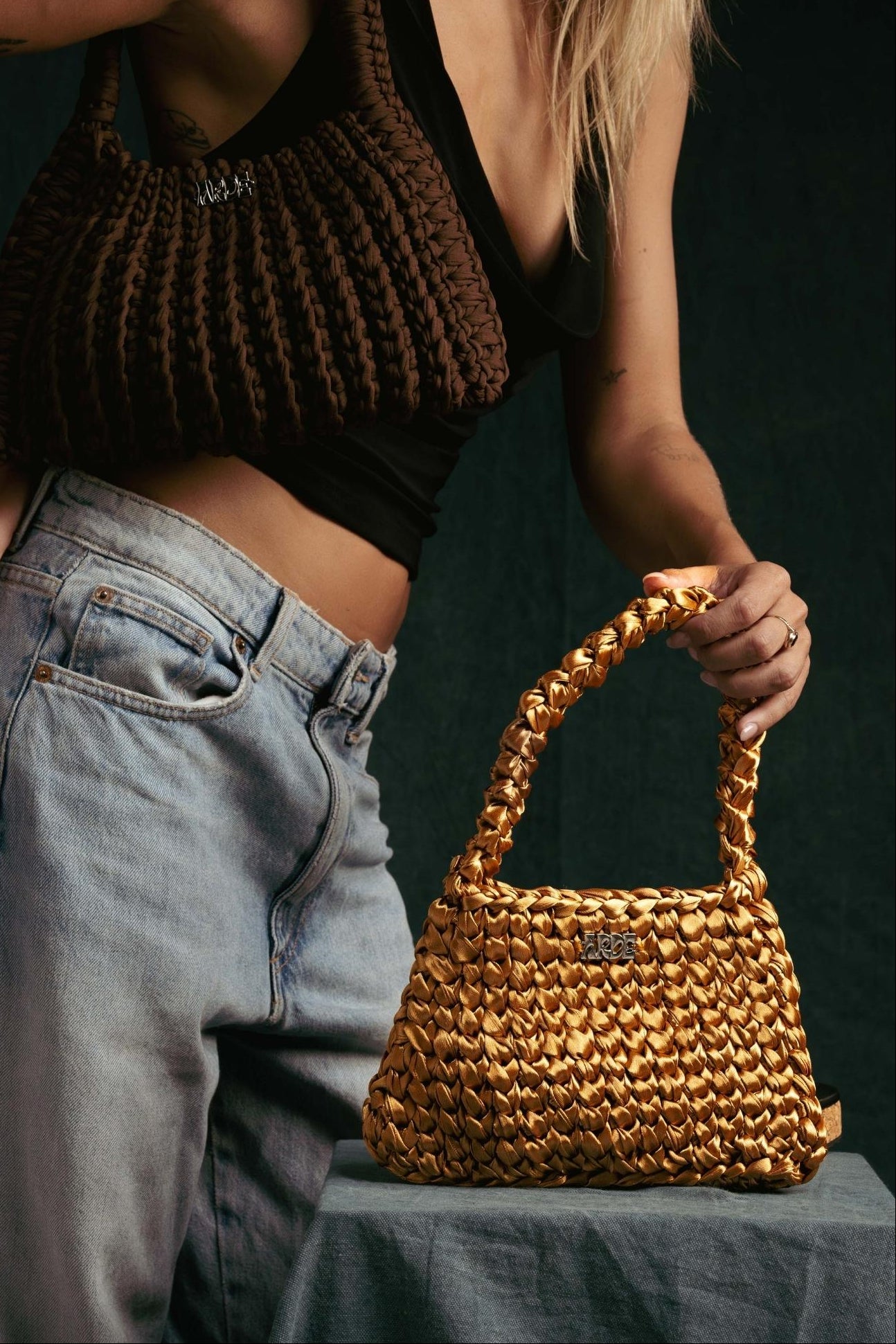 Woman holding a hand-knitted Satin Arde Shoulder Bag in gold with a plain dark-grey background