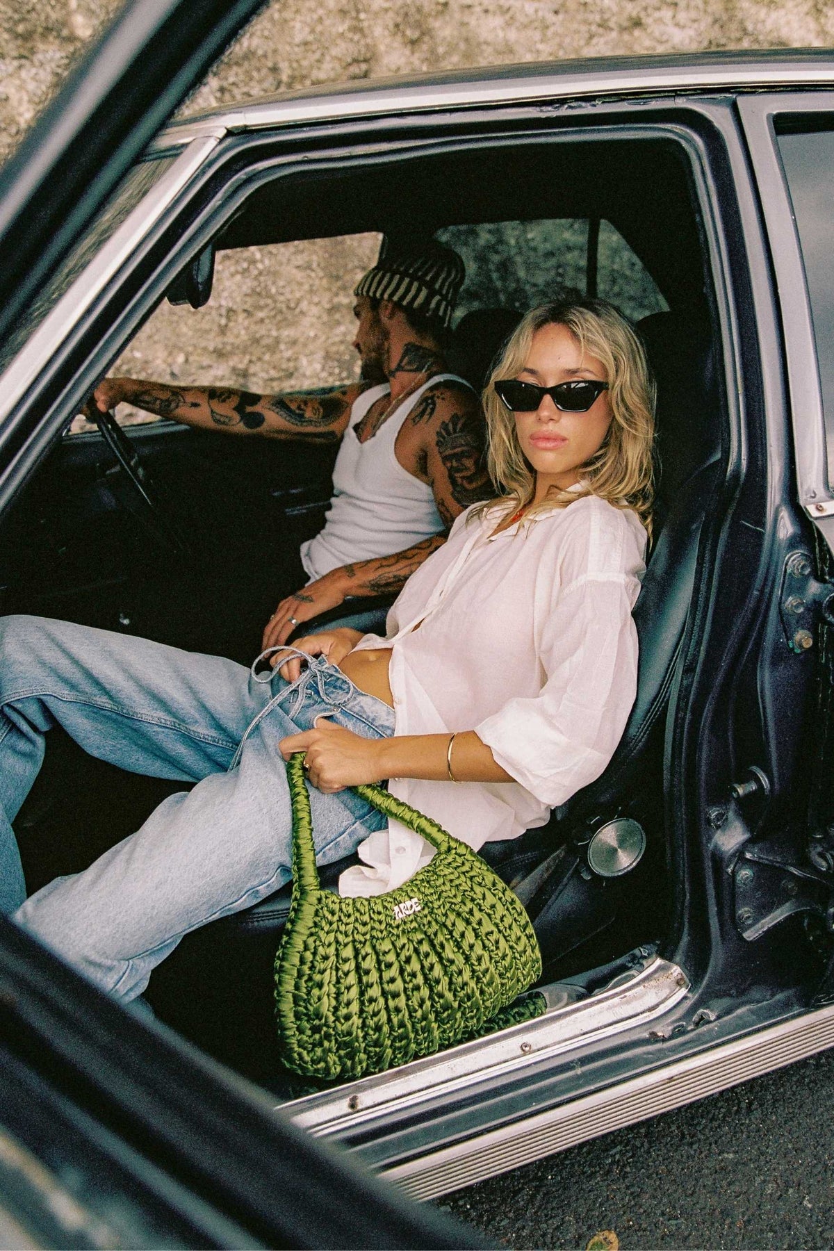 Woman sitting in a classic car holding a green crocheted bag, with a tattooed man in the driver sit