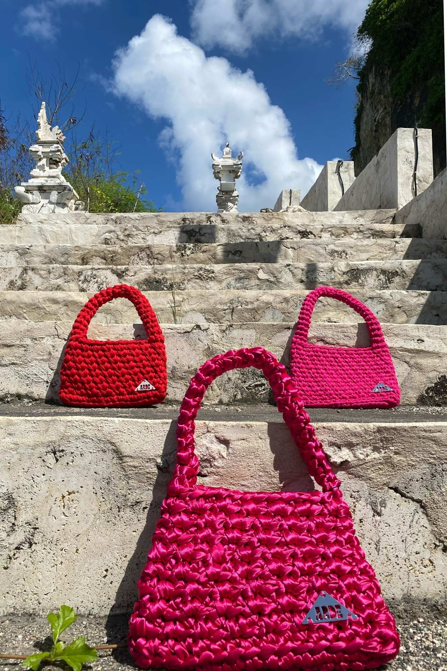 Three pink and red crochet handbags on stone steps with a scenic background