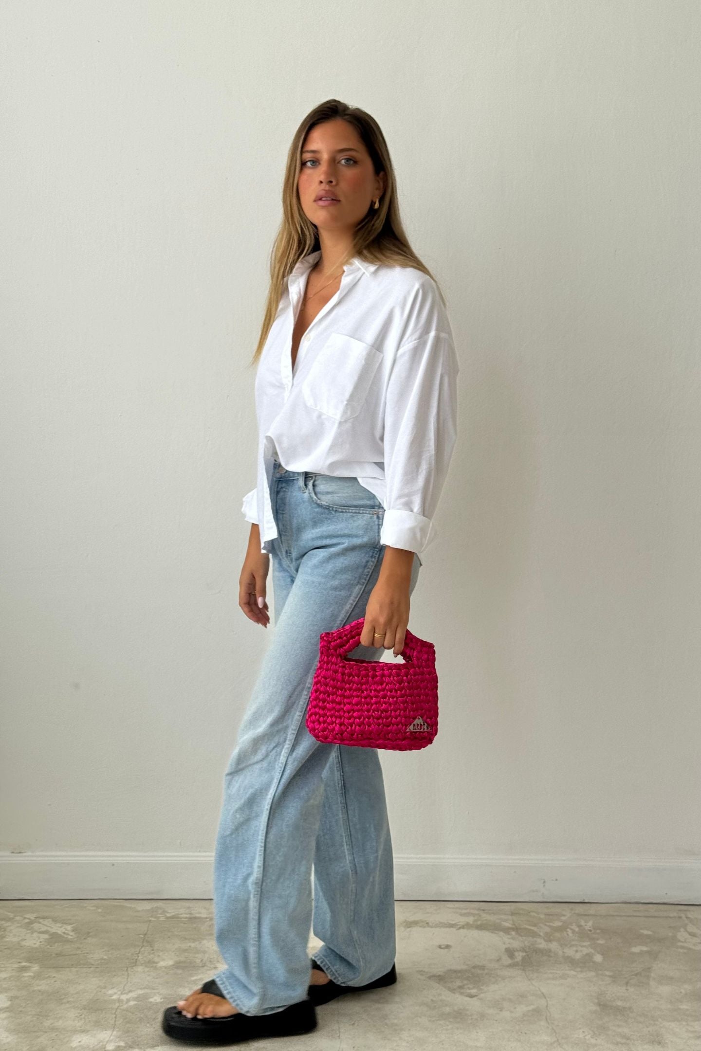 A person standing against a white background, wearing a white long-sleeve top and blue jeans, with a pink satin mini basket handbag.