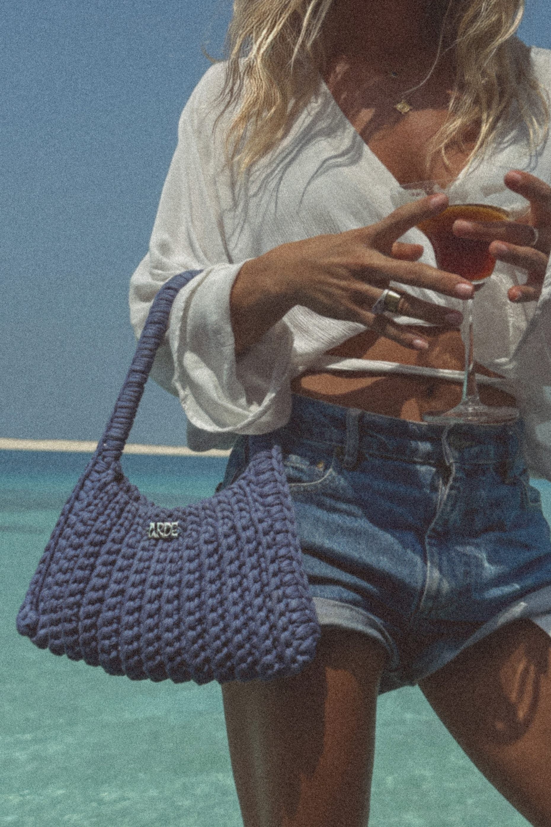 Person holding a glass of red wine and a blue woven handbag by the ocean.