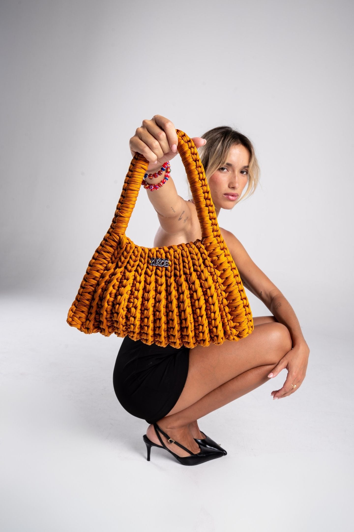 Woman elegant style black mini dress and heels holding a crocheted handmade shoulder bag yellow bag against a white background