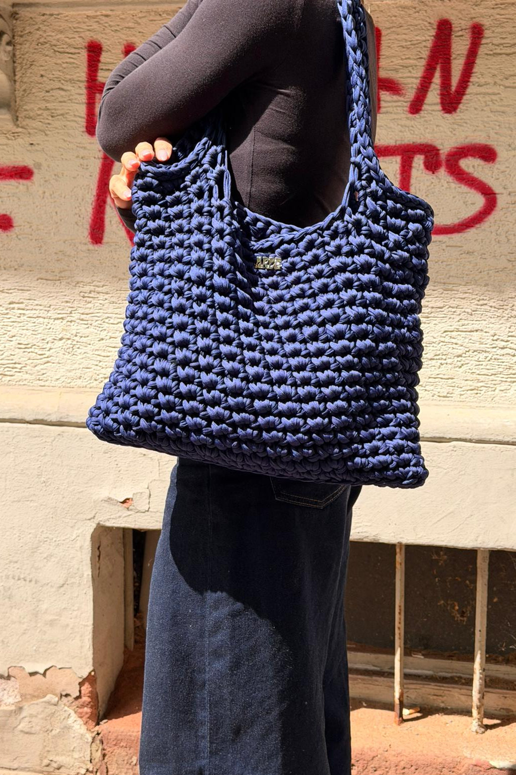 Kenza holding a Navy-colour hand-crocheted tote bag against a wall with graffiti