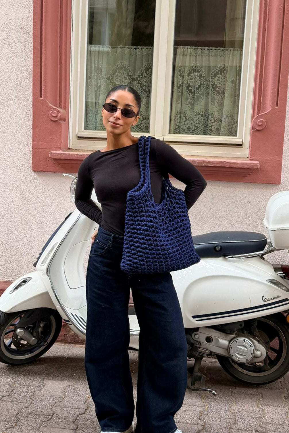 Woman holding a blue knitted bag standing next to a white Vespa scooter.