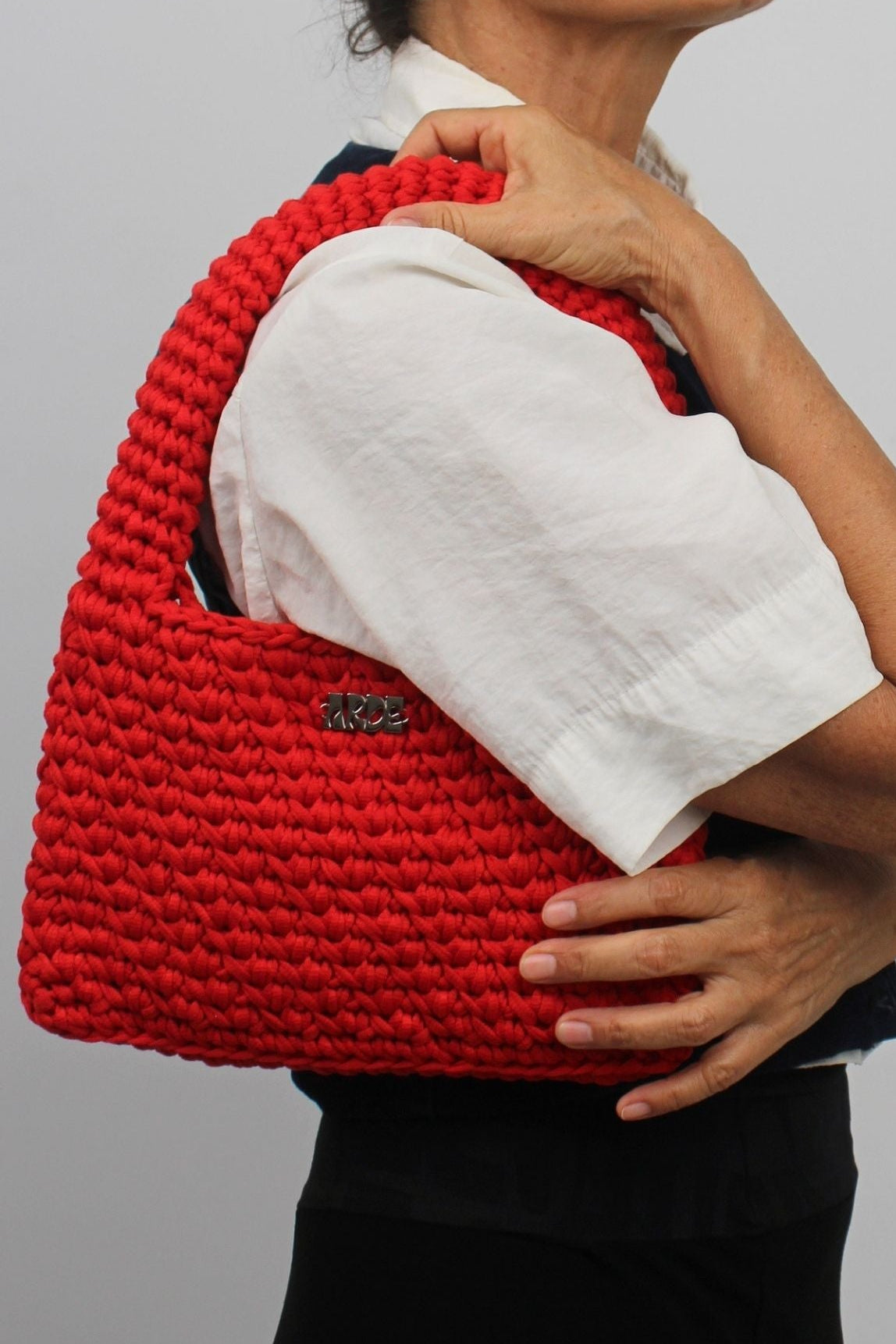 A woman wearing glasses, a white short-sleeve top, and a black skirt carries a large, chunky red woven shoulder bag against her shoulder, photographed against a solid light gray background.