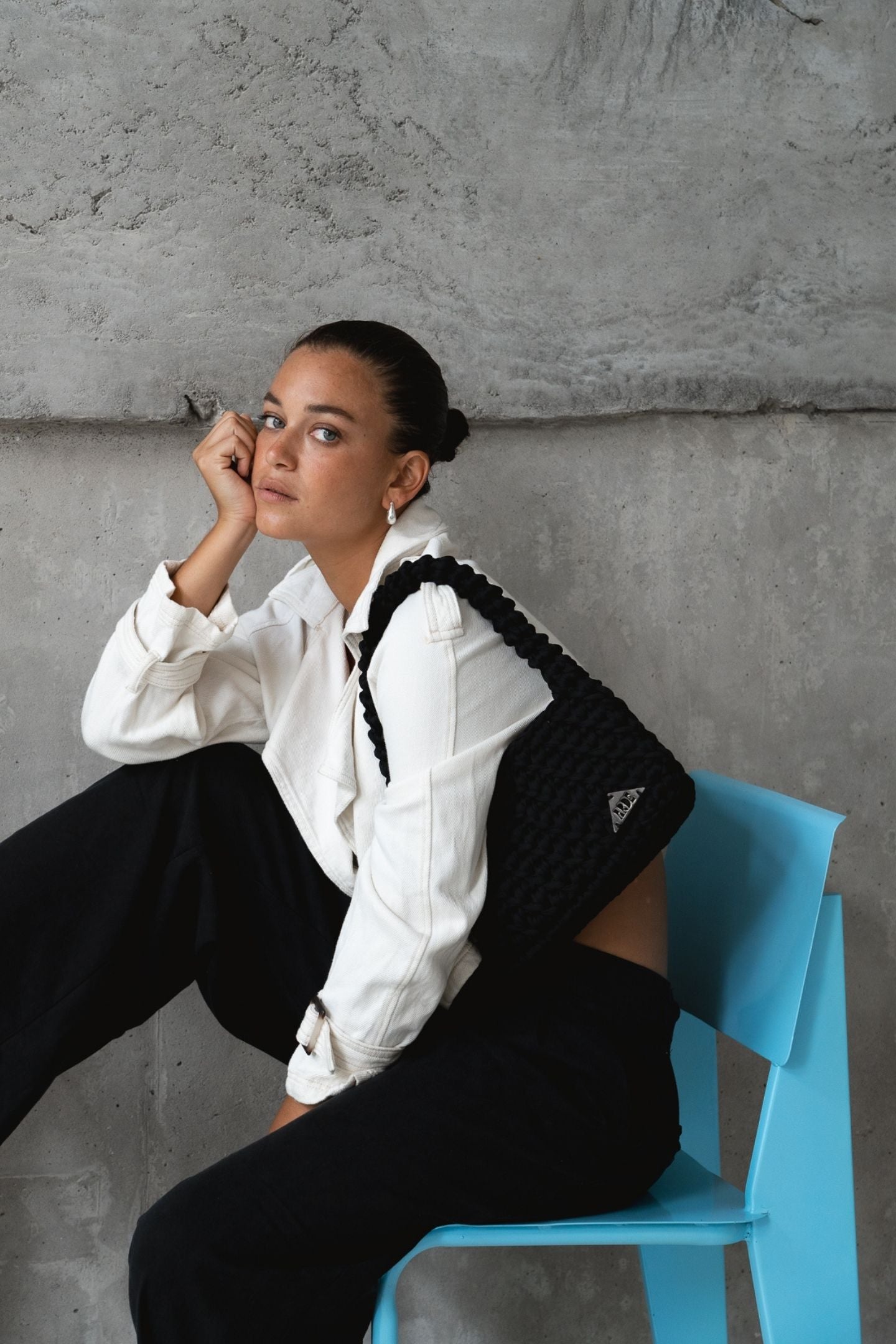 Person sitting on a blue chair against a gray concrete wall wearing a white jacket and a black handcrafted shoulder bag 