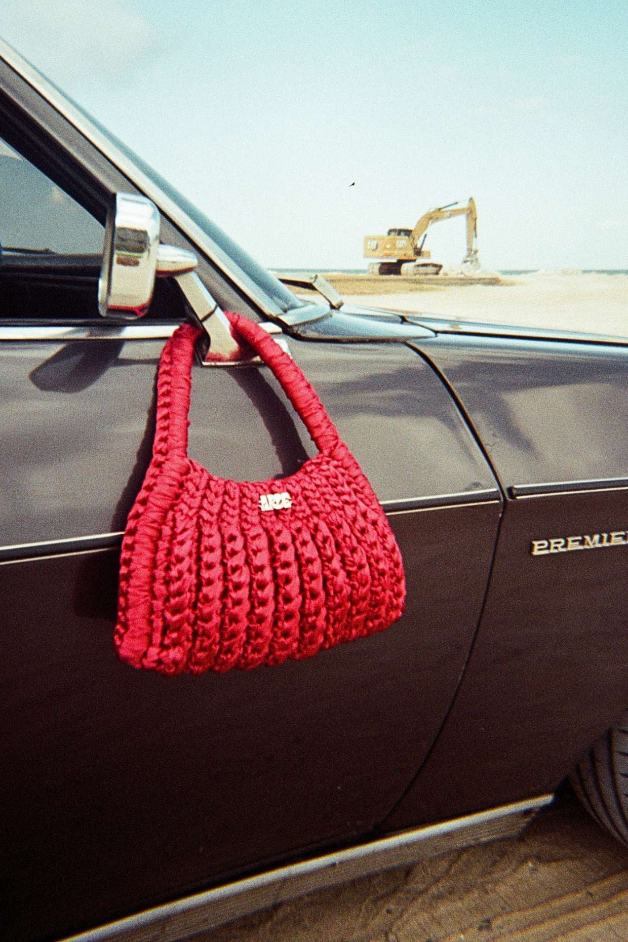Red crocheted sating handbag hanging from a car mirror on a dirt road.