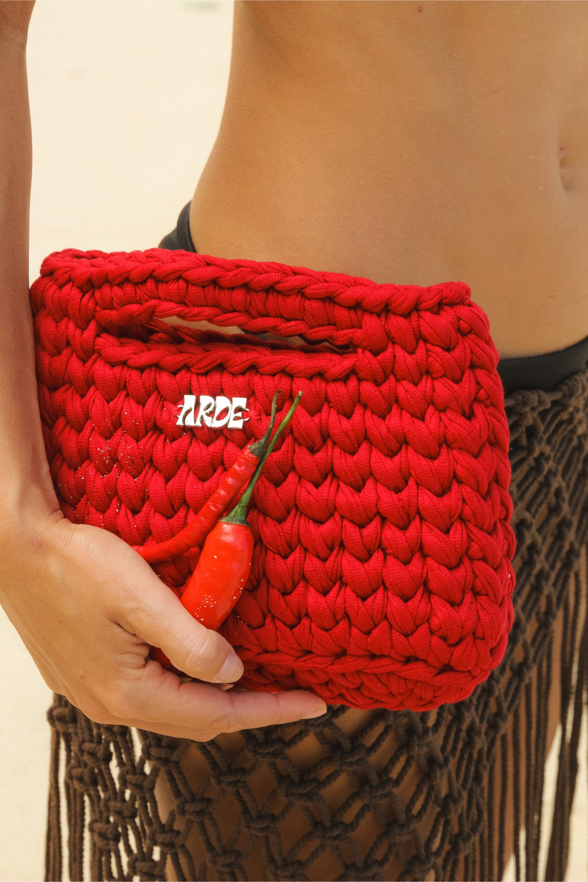 Close-up of ARDE Hand-crocheted Mini Basket Bag with zinc-alloy Logo. Person holding the bag with two red chili peppers.