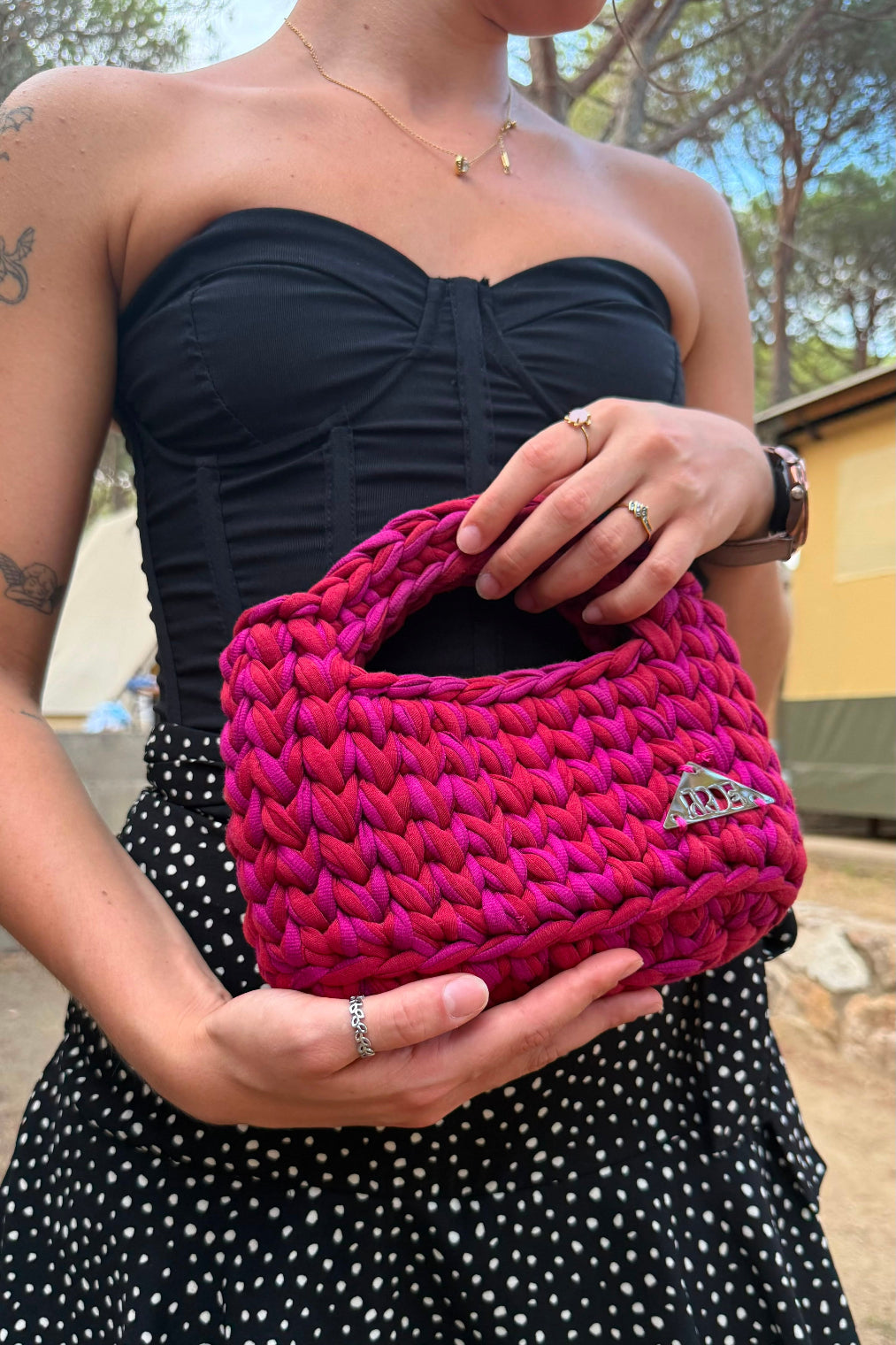 A woman wearing a black strapless top and black-and-white polka dot skirt holds a bright pink chunky knit shoulder bag with a small silver triangular logo plaque.