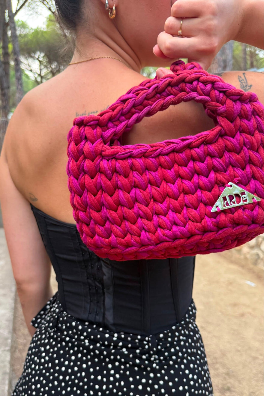 A close-up lifestyle shot of a woman from behind wearing a black strapless corset top and a black-and-white polka dot skirt, carrying a vibrant hot pink braided shoulder bag with a silver-tone triangular logo plaque.