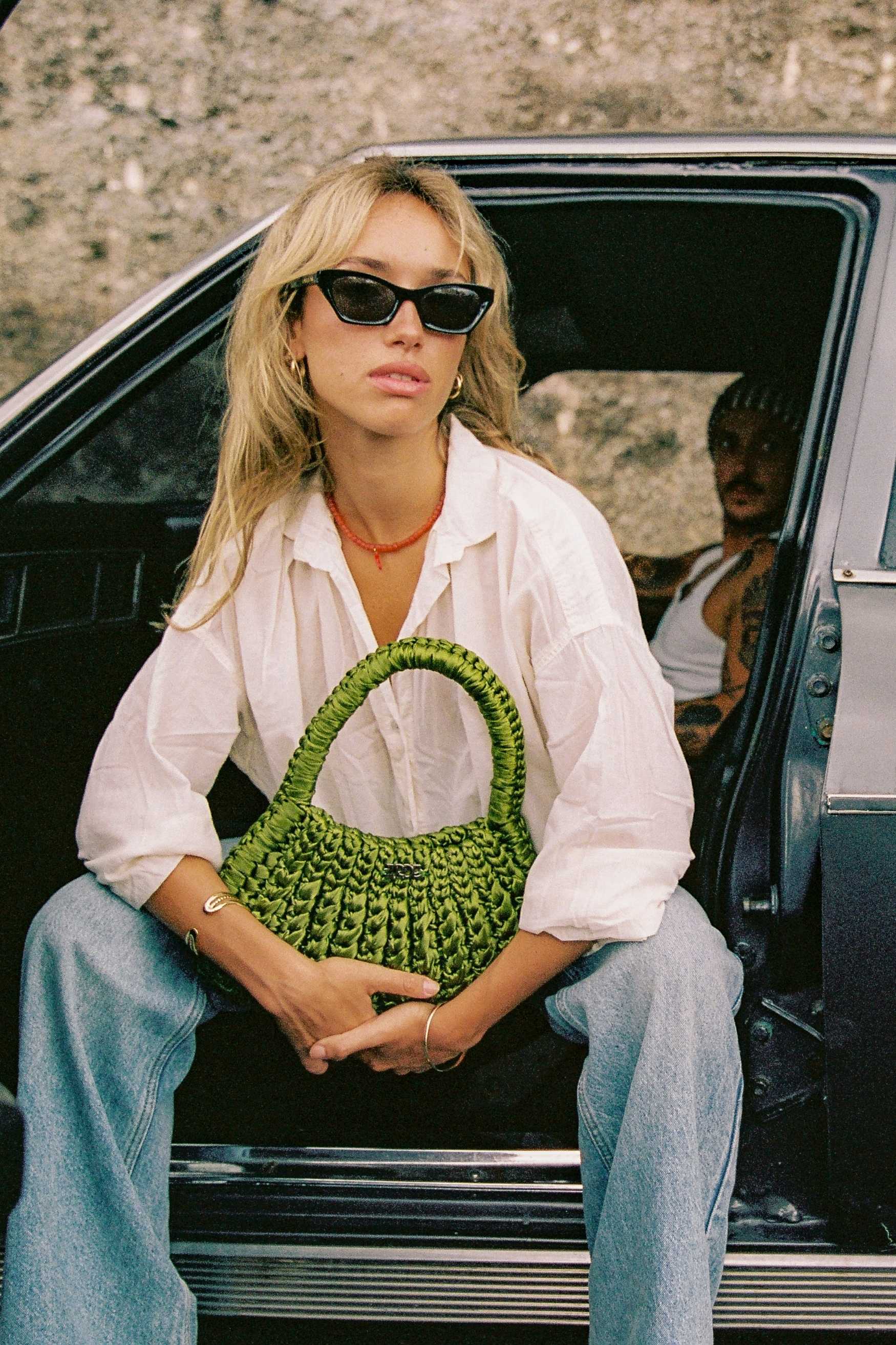 Person sitting in a car holding a green woven handbag, wearing sunglasses and a white shirt.