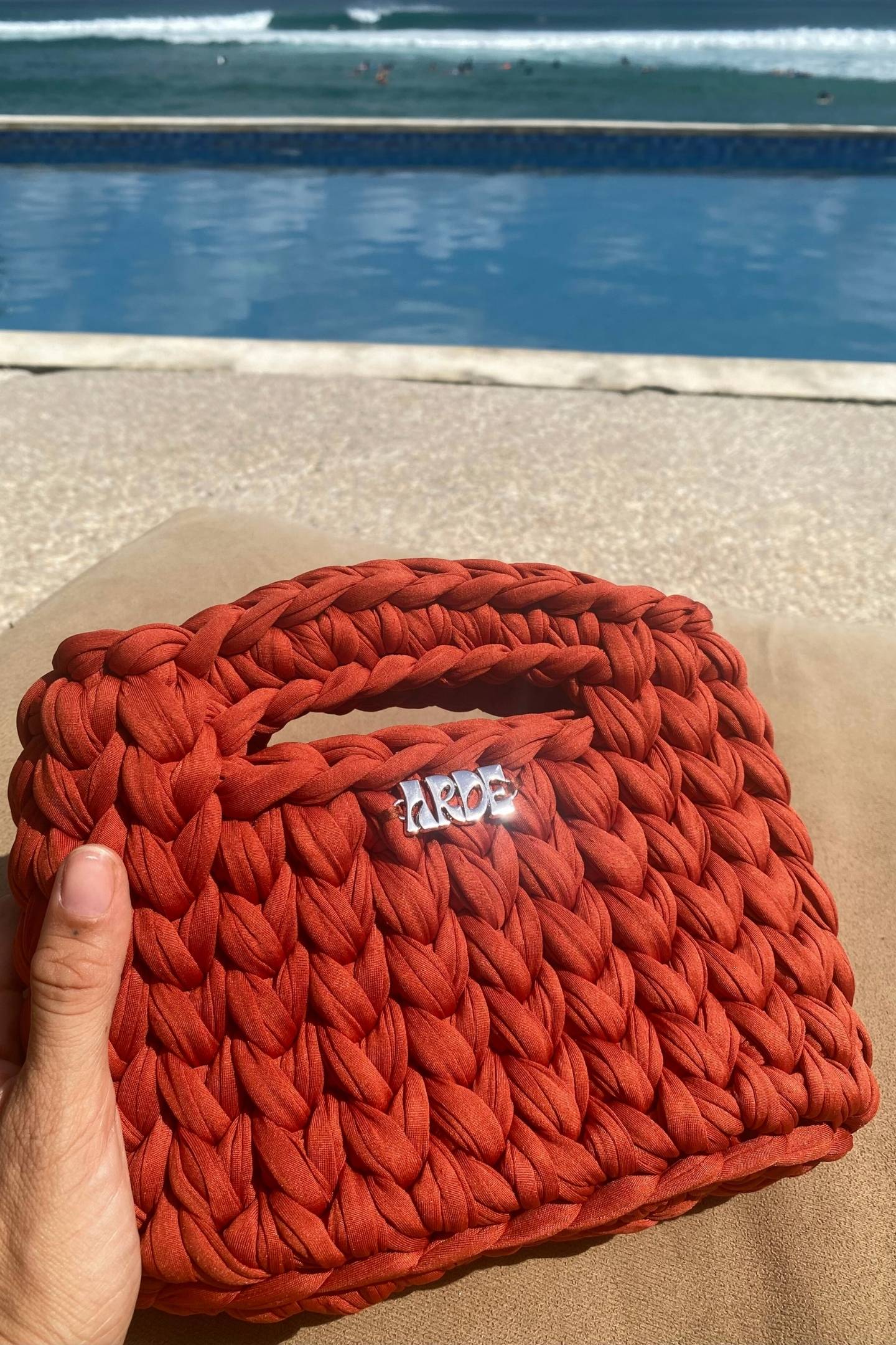 Dark amber colour woven handbag with a visible brand logo held by a hand against a poolside background.